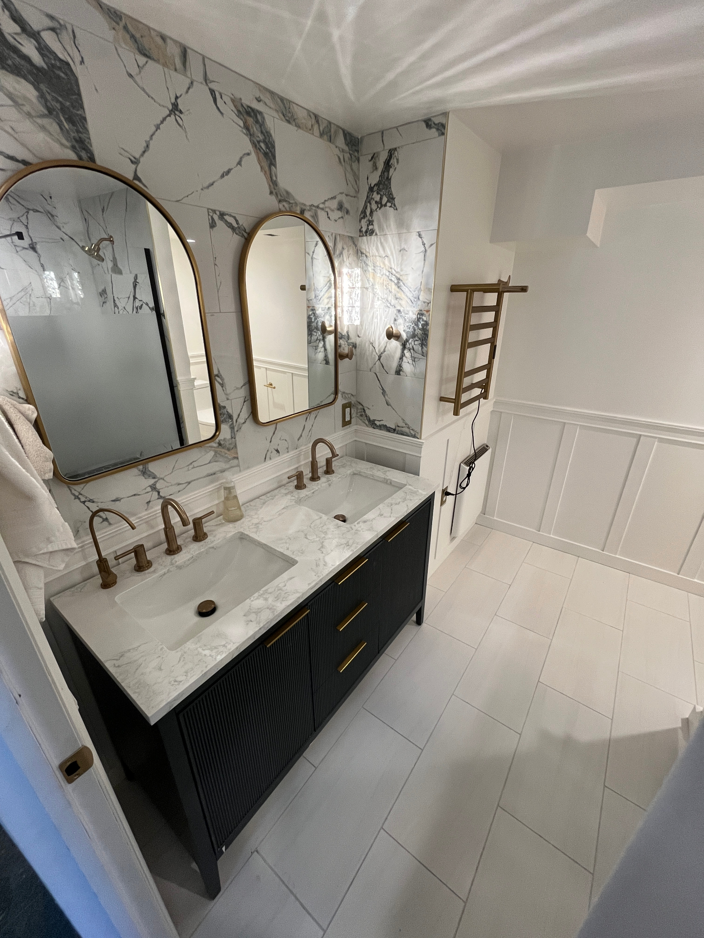 Finished marble bathroom with black fluted double vanity, brass arched mirrors, and brass towel warmer
