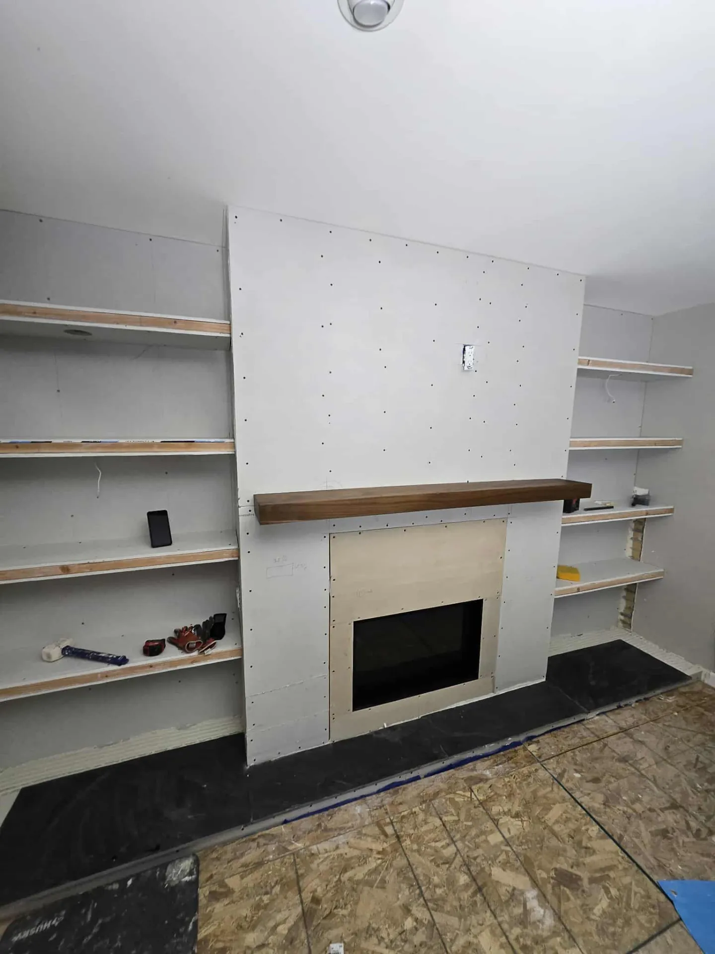 Fireplace feature wall during construction with drywall, framed shelf ledgers, and walnut mantel set